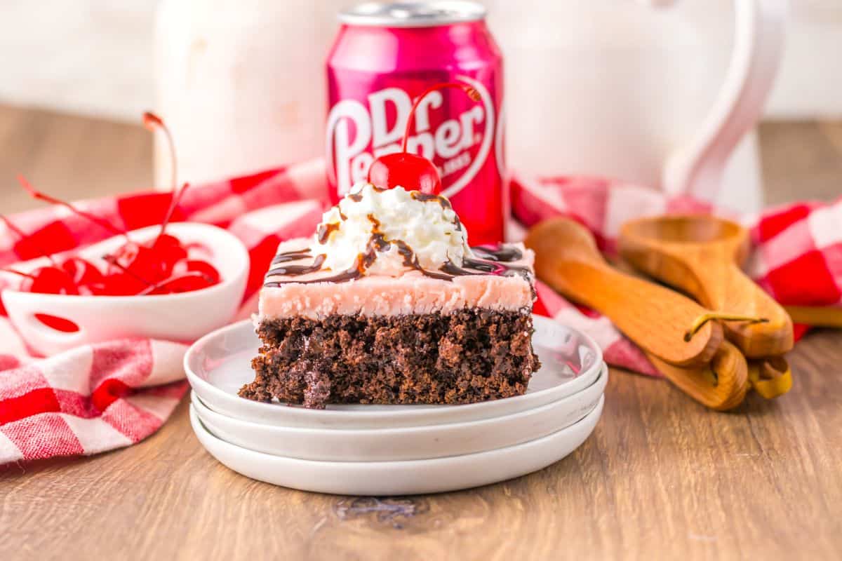 A slice of chocolate cake with pink frosting, whipped cream, chocolate drizzle, and a cherry on top sits on a plate beside a can of Dr Pepper and a bowl of cherries.