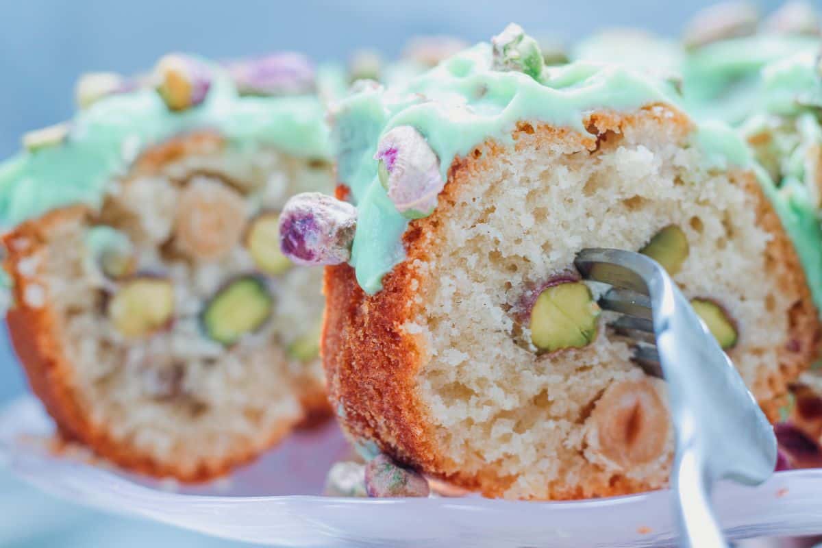 A close-up of a pistachio cake with green icing, topped and filled with whole pistachios, with a fork cutting a slice.