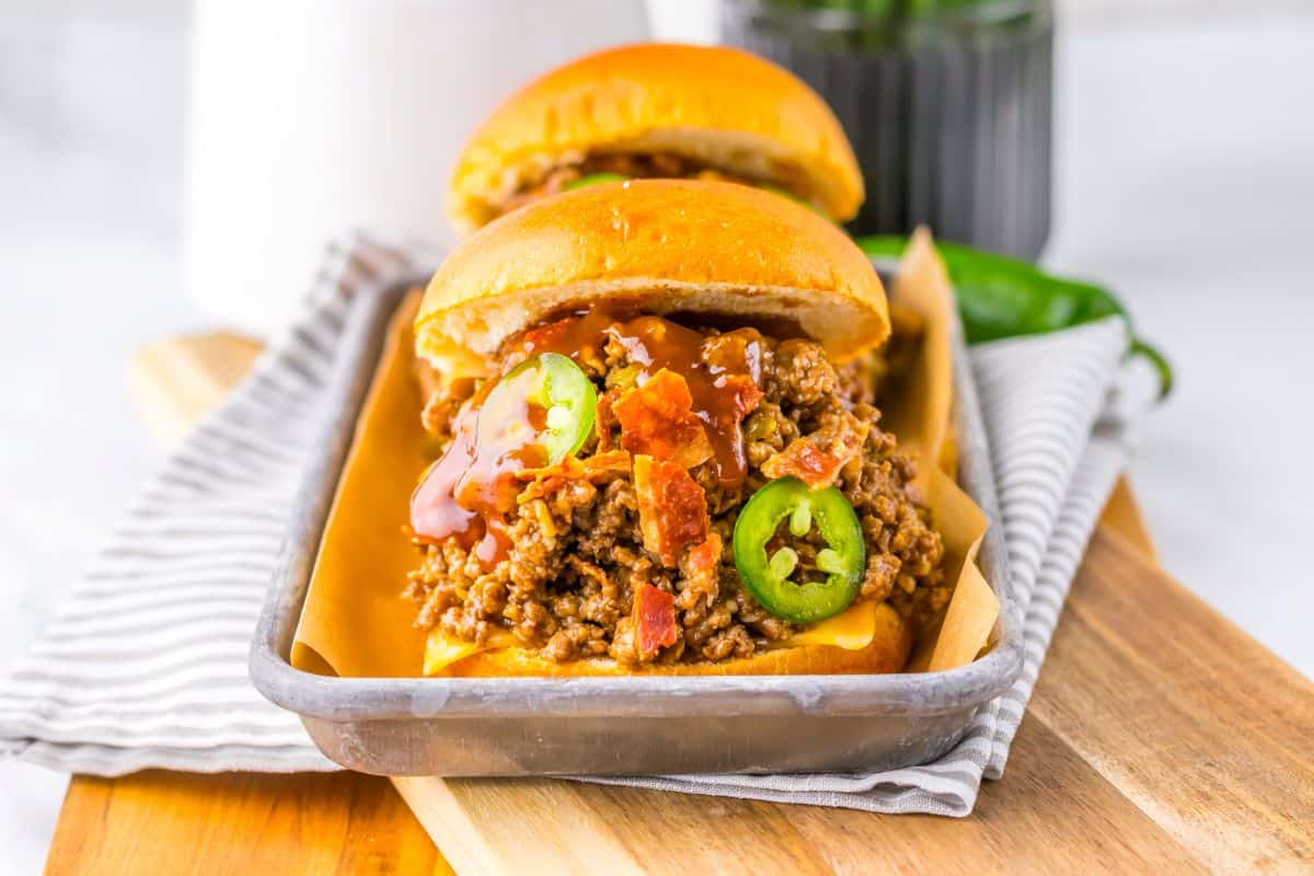 Two sloppy joe sandwiches topped with sliced jalape&ntilde;os and sauce are served on a metal tray lined with parchment paper, with a striped napkin underneath.