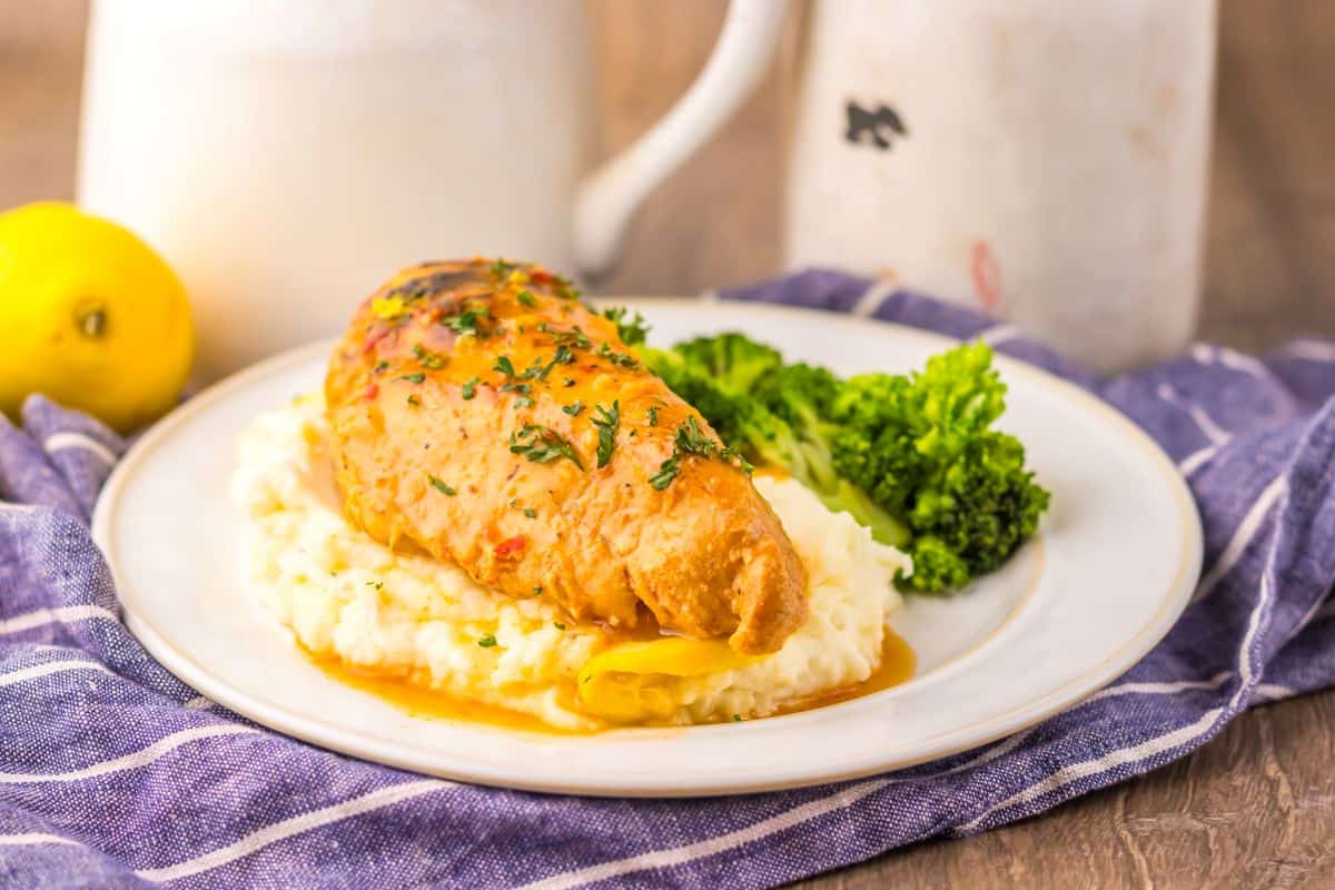 A stuffed chicken breast topped with herbs is served on mashed potatoes with a side of steamed broccoli on a white plate.