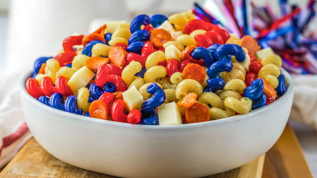 A bowl of tri-color pasta salad featuring red, blue, and plain pasta spirals, mixed with small cubes of cheese and slices of pepperoni.