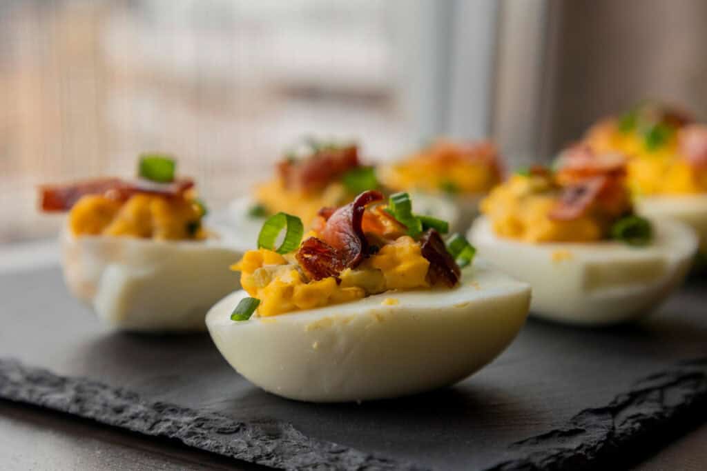 Close-up of deviled eggs topped with chopped green onions and bacon pieces, arranged on a dark slate platter near a window.