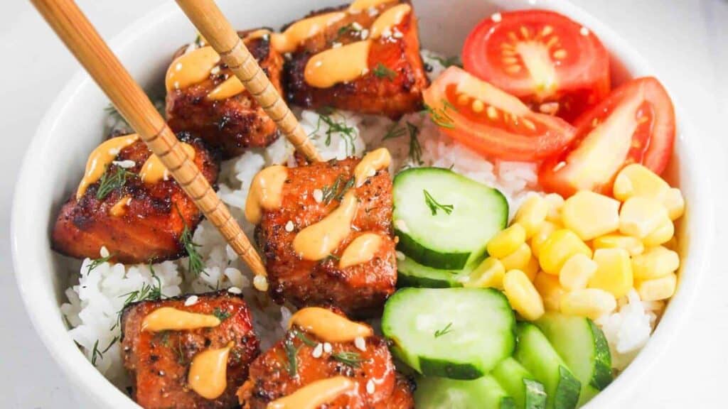 A bowl of white rice topped with glazed salmon cubes, sliced cucumbers, corn, tomato wedges, and a drizzle of orange sauce offers a fresh take on global recipes that beat the same old rotation&mdash;complete with chopsticks picking up a salmon piece.