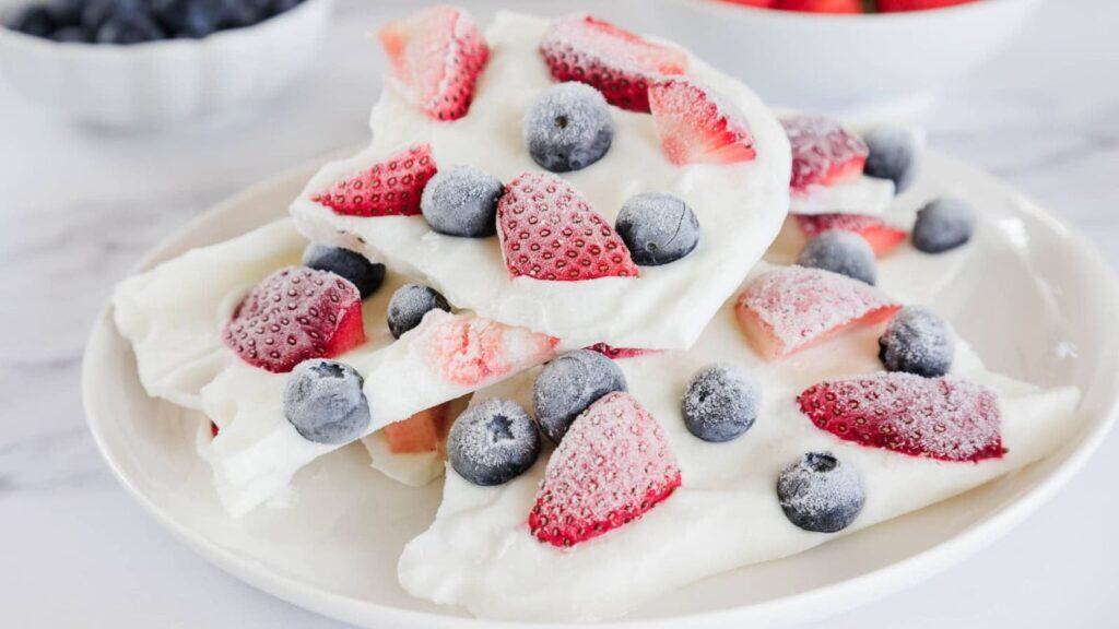 A plate with pieces of frozen yogurt bark topped with sliced strawberries and whole blueberries, with bowls of berries in the background.