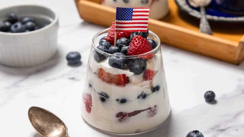 A glass of yogurt parfait with strawberries and blueberries, topped with a small American flag, sits on a marble surface beside a spoon and a bowl of blueberries.