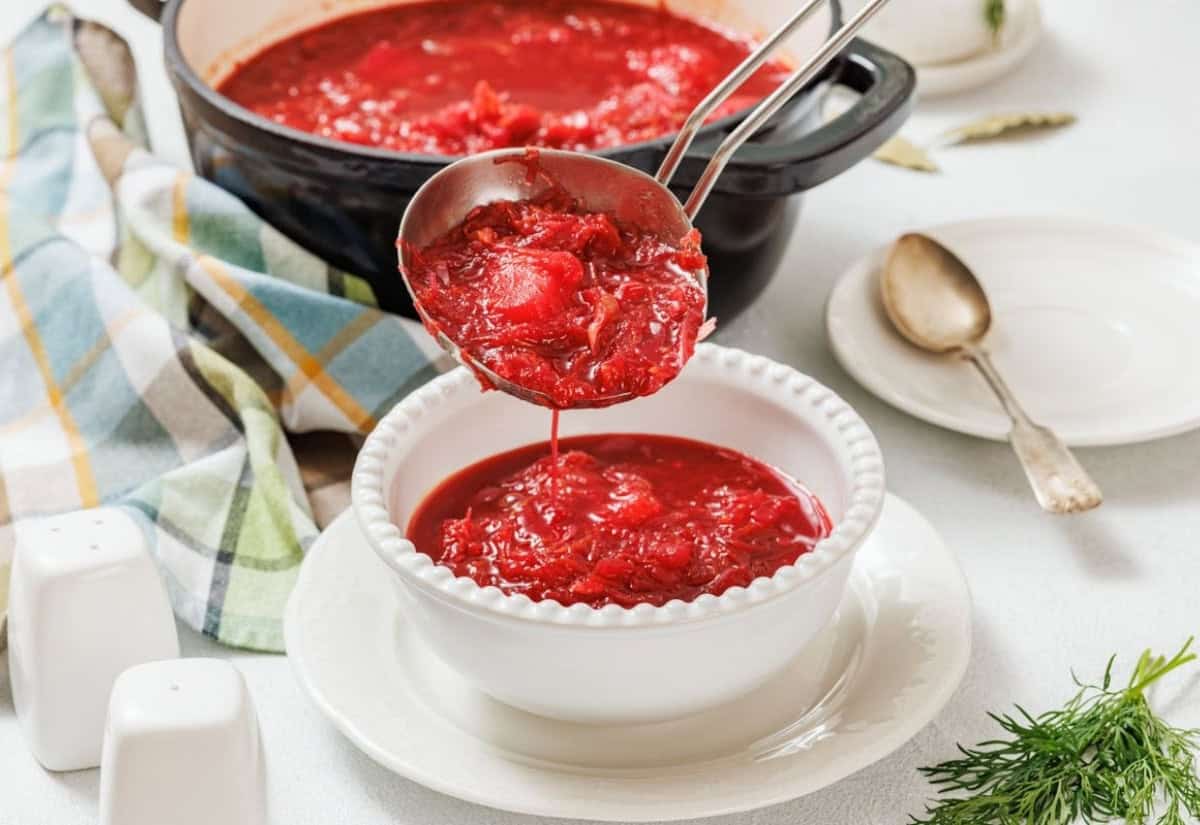 A ladle pours vibrant red borscht into a white bowl, set on a table with a striped cloth and dill garnish. A rustic, cozy kitchen vibe is conveyed.