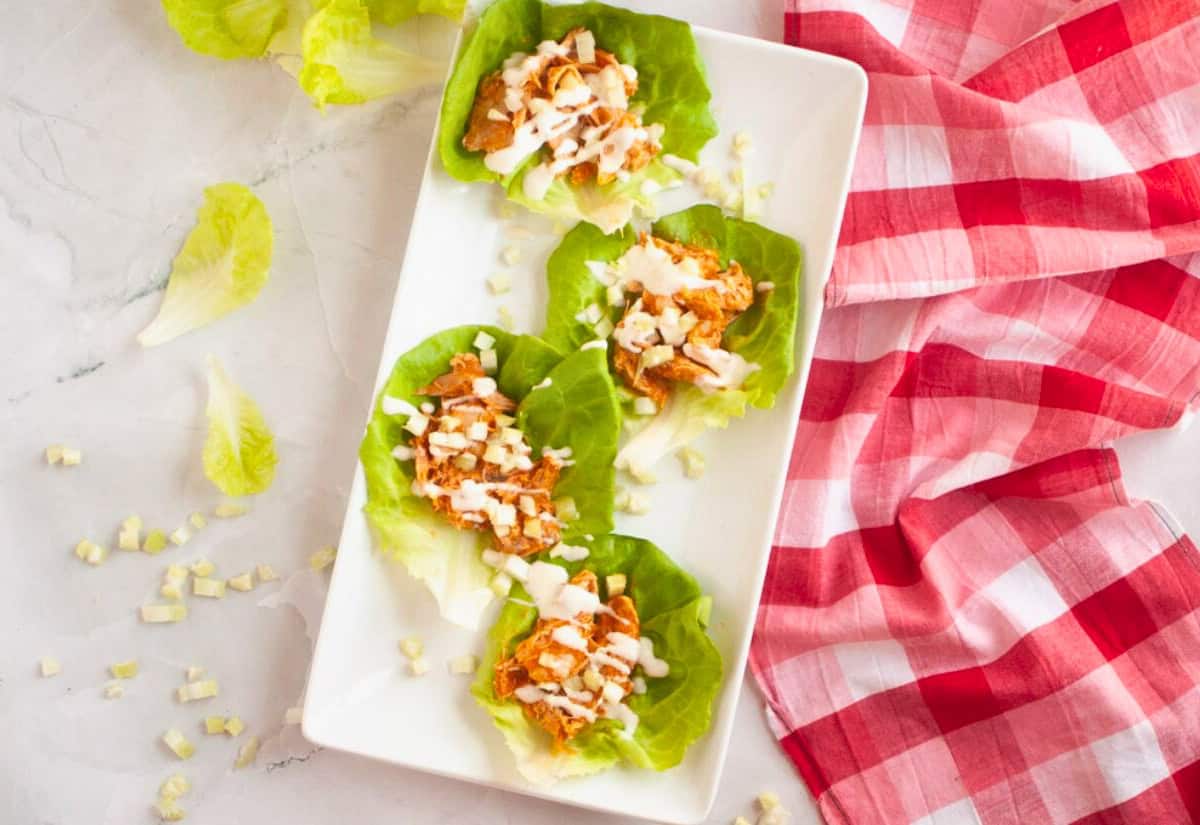 Four lettuce wraps filled with shredded chicken and dressing on a white plate, surrounded by fresh lettuce and a red checkered cloth. Cream dressing in a small bowl nearby.