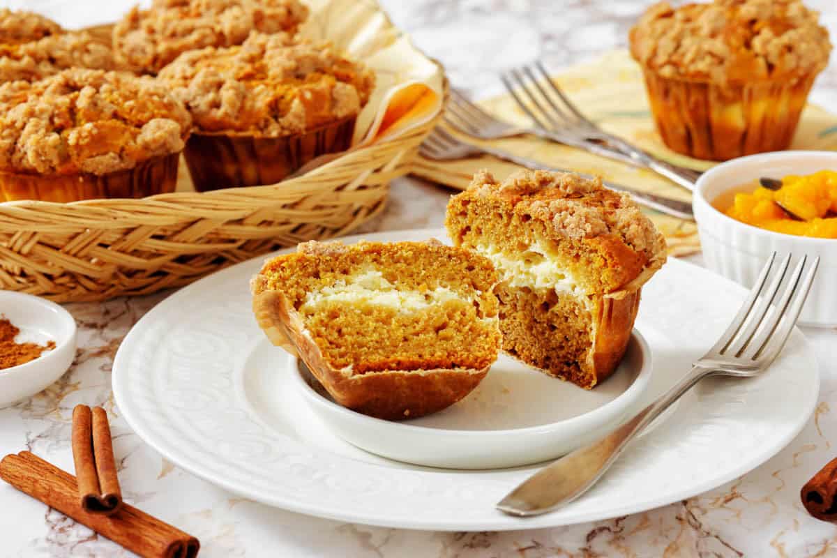 A Pumpkin Cream Cheese Muffins cut in half on a plate reveals a creamy filling, with more muffins in a basket and a bowl of diced fruit in the background.