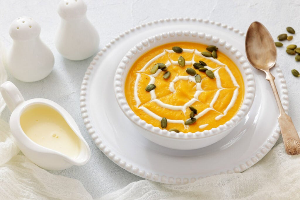 A bowl of creamy pumpkin soup garnished with cream and pumpkin seeds, served on a white plate with a gold spoon, next to a small pitcher and salt and pepper shakers.