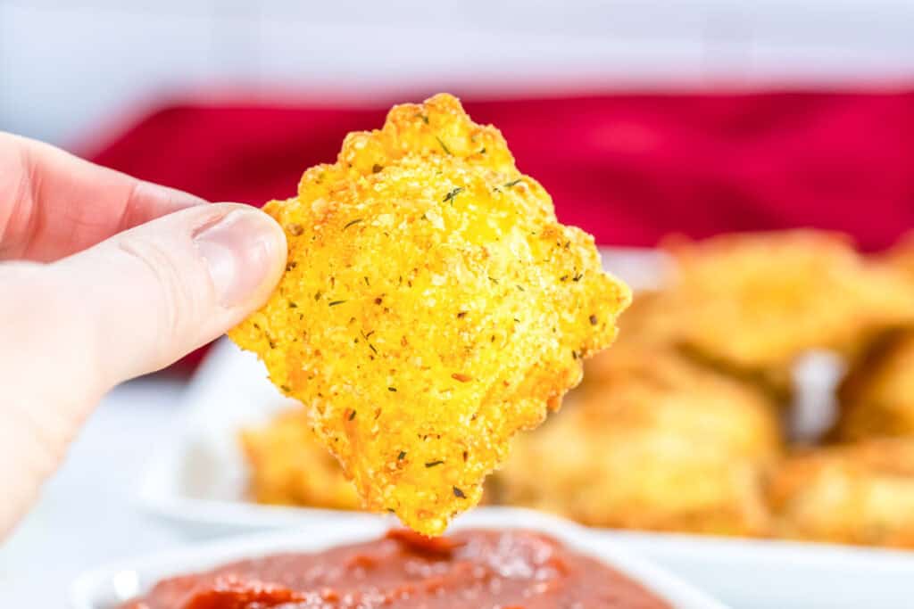 A hand holds a piece of breaded, fried ravioli above a bowl of marinara sauce, with more ravioli on a plate in the background.