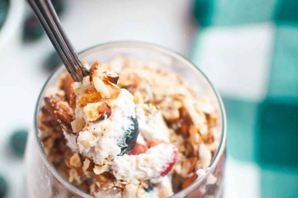 A close-up view of a berry trifle with whipped cream, berries, and chopped nuts in a glass, with a spoon inserted.