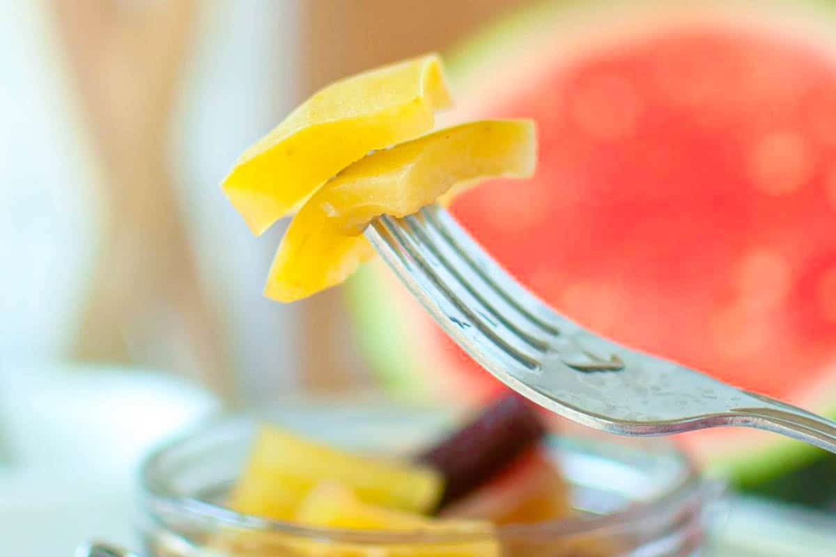 A close-up of a fork holding several slices of pickled watermelon with a blurred background, including part of a watermelon.