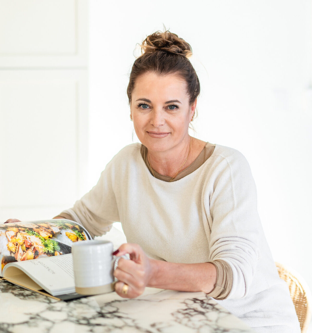 A woman sits at a marble table holding a mug, with an open cookbook in front of her, in a bright kitchen.