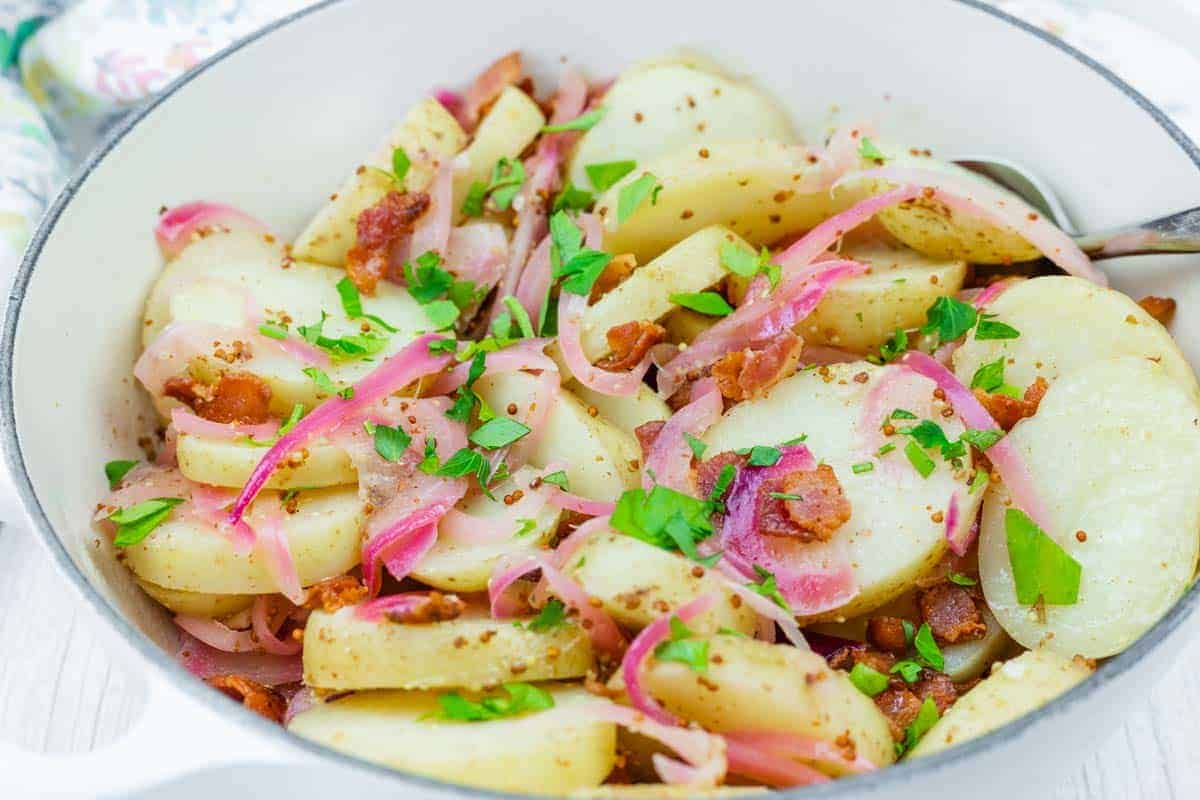 German Potato Salad with red onions, and bacon pieces, garnished with chopped parsley are served in a white dish with a spoon.
