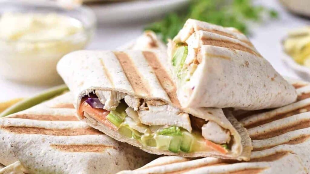 Grilled wraps filled with sliced chicken, lettuce, cucumber, and other vegetables, stacked on a plate.