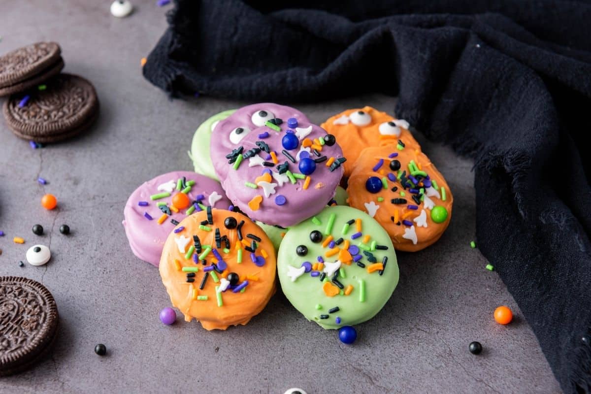 A group of Halloween Oreo Cookies coated in green, orange, and purple, decorated with sprinkles and candy eyes, sits on a gray surface next to a black cloth.