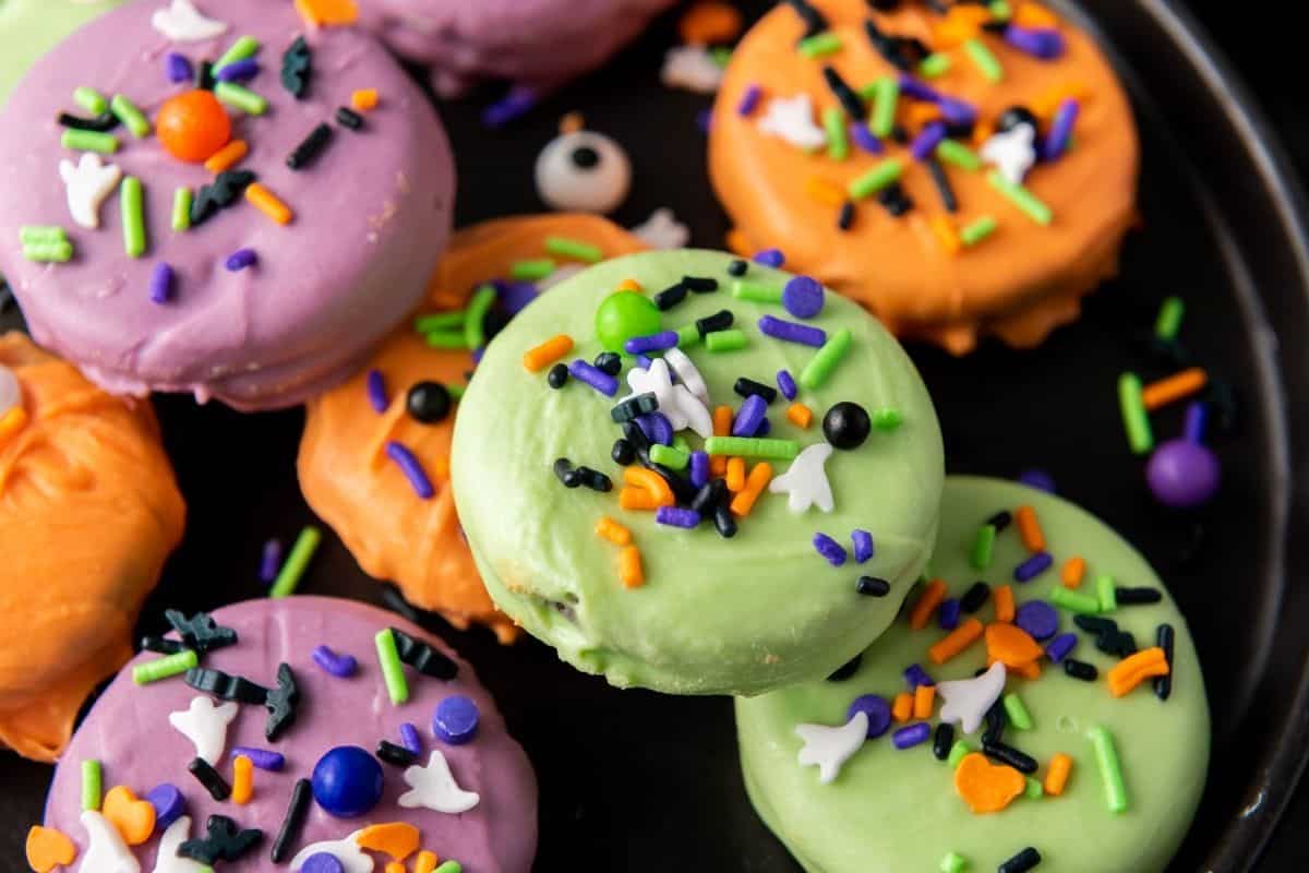 Colorful cookies decorated with green, orange, and purple icing and topped with assorted Halloween-themed sprinkles on a black plate.