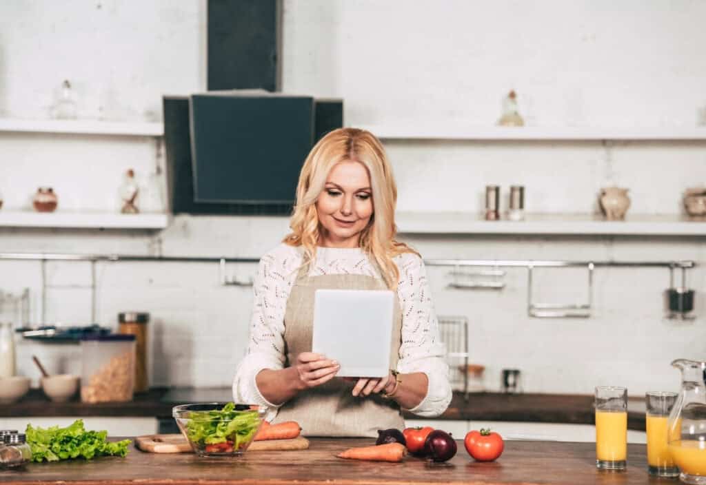 A woman wearing an apron uses a tablet in a kitchen, surrounded by fresh vegetables, salad, and glasses of juice on the counter, as she focuses on meal planning.