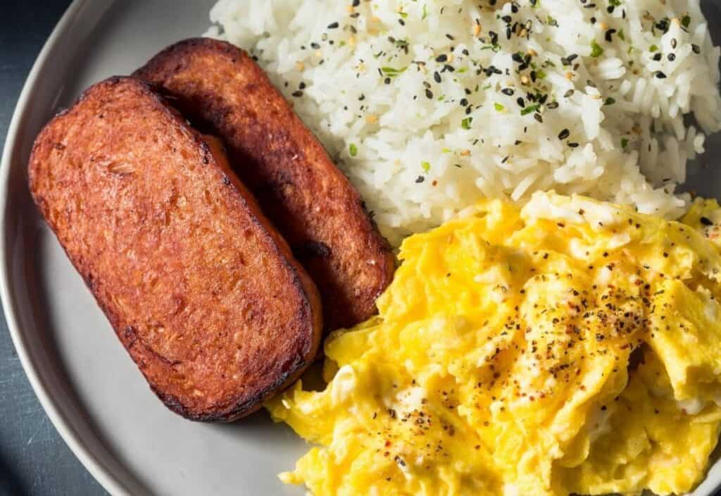 A plate with two slices of cooked Spam, a serving of scrambled eggs with black pepper, and a portion of white rice garnished with seasoning.
