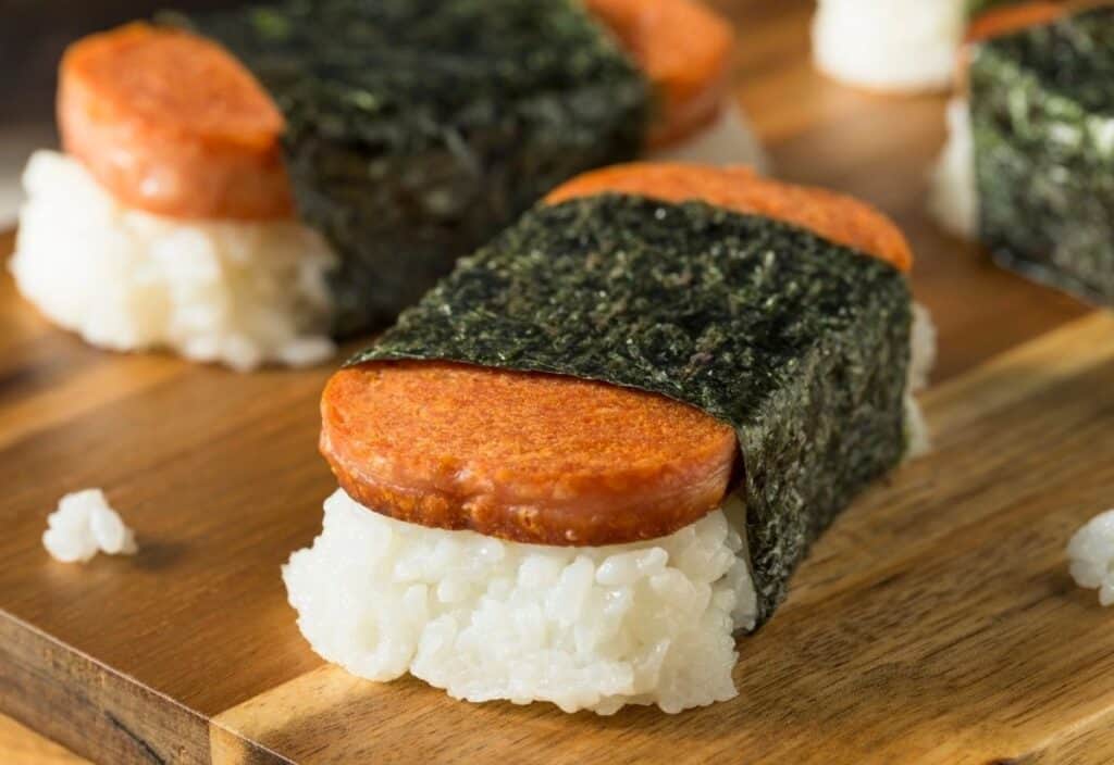 Slices of spam on top of white rice, wrapped with a strip of seaweed, arranged on a wooden surface.