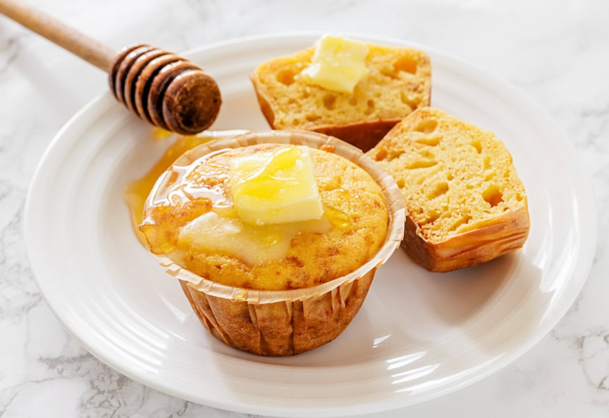 A freshly baked corn muffin on a white plate, topped with melting butter and honey. Nearby, a split muffin half also has butter, exuding warmth and comfort.