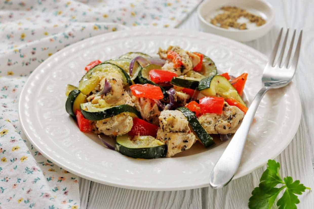 A white plate with a serving of Italian Herbed Chicken & baked foil packs with a fork on the side and a small bowl of seasoning in the background.