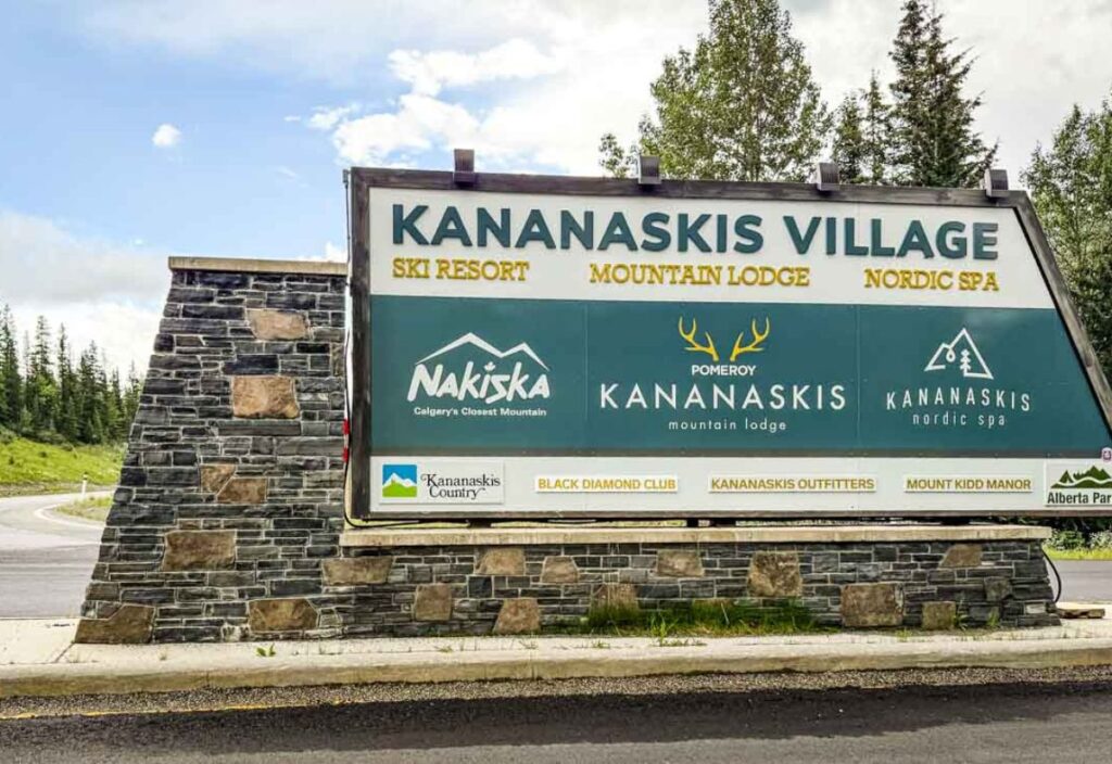 Large sign for Kananaskis Village advertising a ski resort, mountain lodge, and nordic spa, with kananaskis-themed logos on a stone base, set near the road and surrounded by trees.