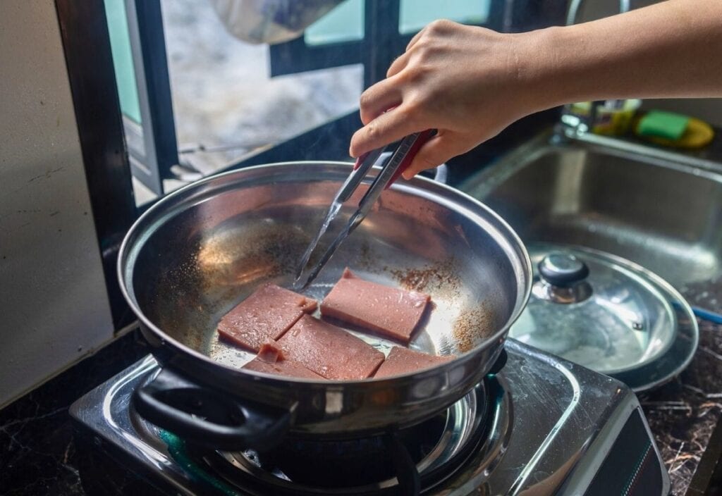 A person cooks slices of luncheon meat in a stainless steel pan on a gas stove, using tongs to turn the pieces.