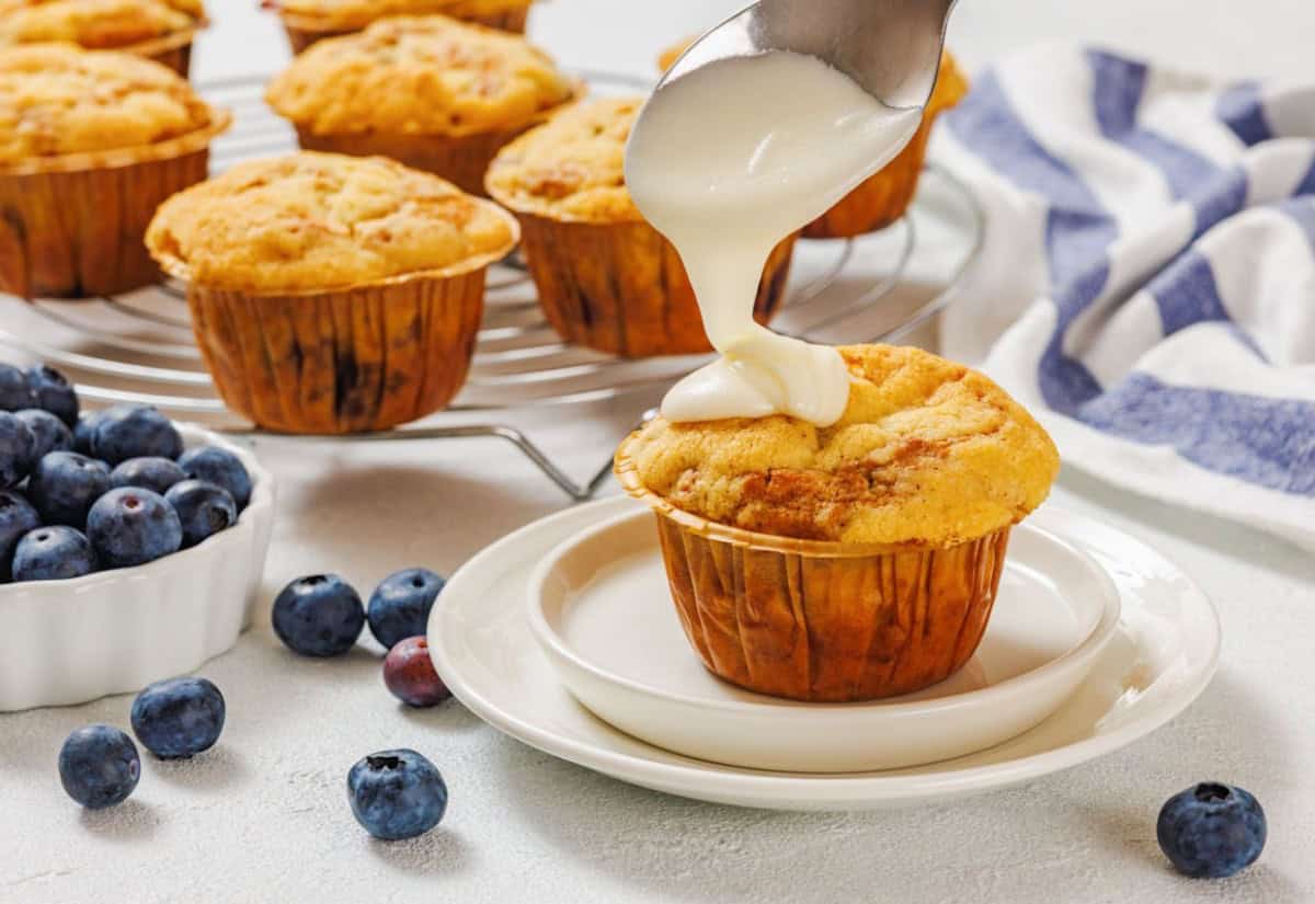 Golden muffins on a wire rack, with one on a plate being drizzled with white icing. Fresh blueberries scattered nearby, adding a fresh touch.