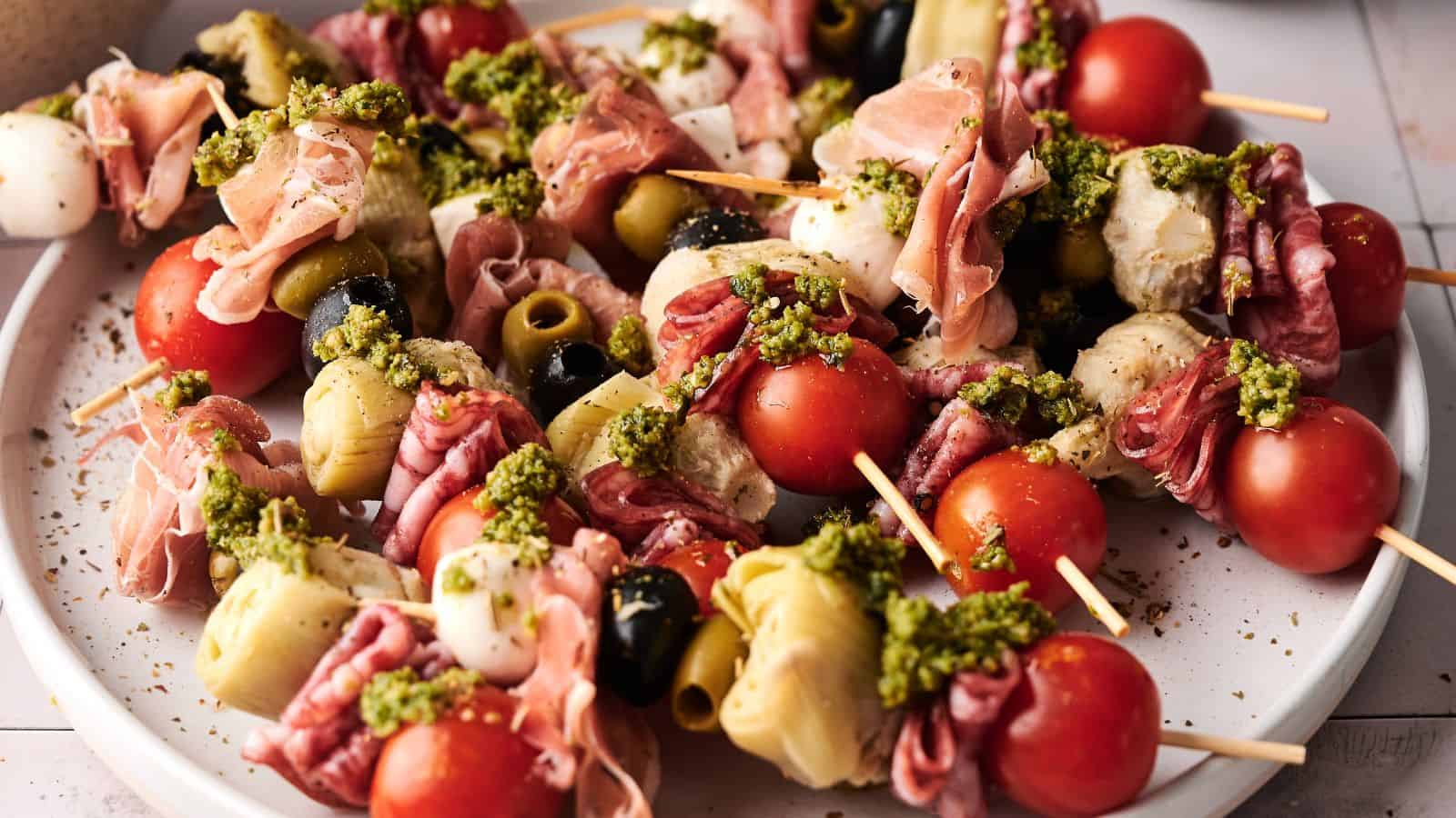 A plate of antipasto skewers with cherry tomatoes, mozzarella, olives, artichoke hearts, cured meats, and a drizzle of green pesto.
