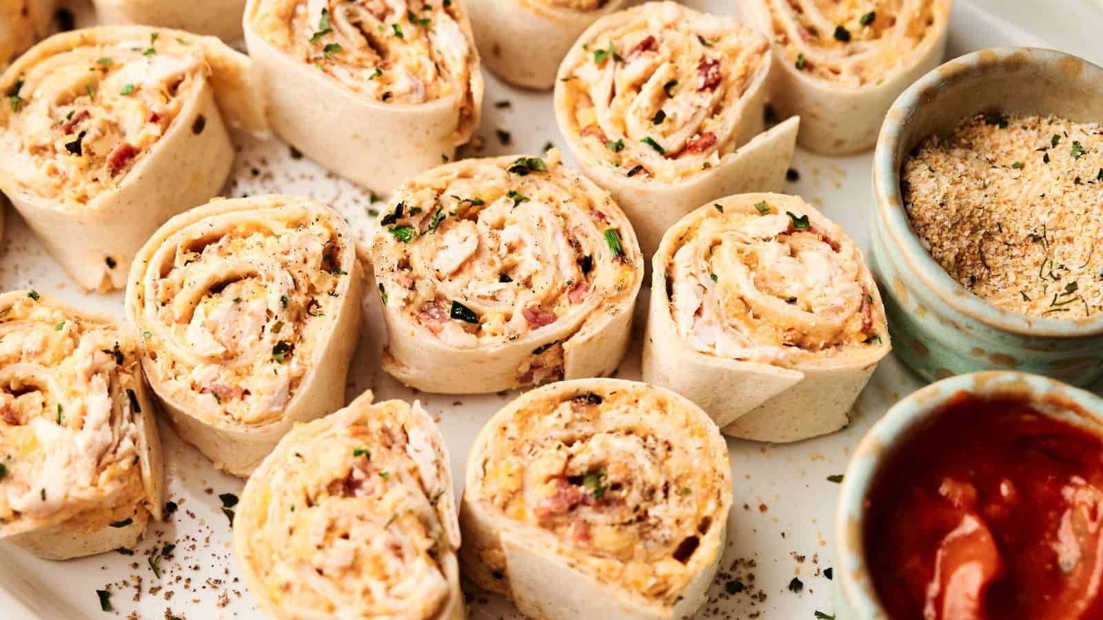 A plate of tortilla pinwheels filled with a creamy mixture, garnished with herbs, served with bowls of salsa and breadcrumbs.