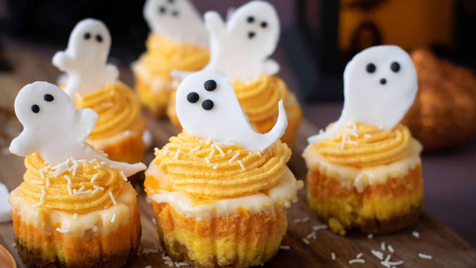 Mini cupcakes with yellow frosting and white ghost-shaped toppers decorated with black eyes, arranged on a wooden surface with sprinkles.