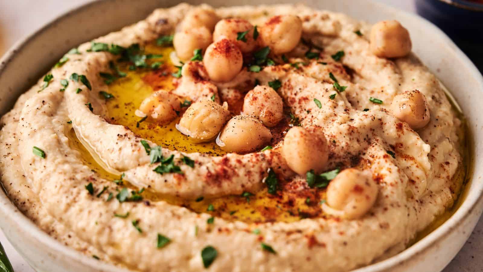 A bowl of hummus garnished with whole chickpeas, chopped parsley, paprika, and a drizzle of olive oil.