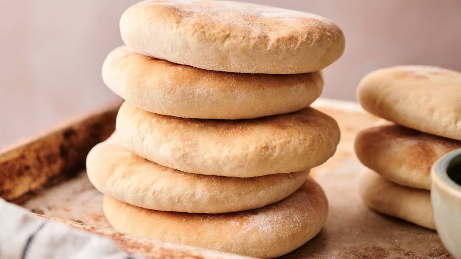 A stack of six round, golden-brown pita breads sits on a baking tray, with more pita breads visible to the side.
