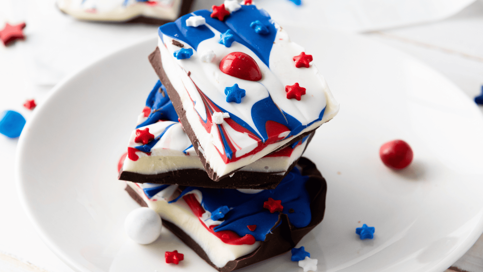 Three pieces of chocolate bark with red, white, and blue swirled topping and star-shaped sprinkles stacked on a white plate.