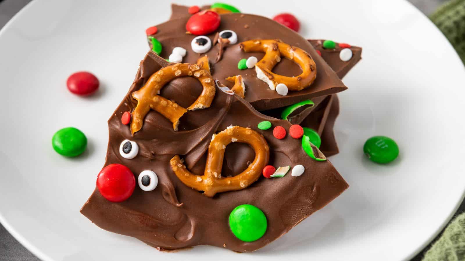 Chocolate bark topped with mini pretzels, red and green candy-coated chocolates, candy eyes, and round sprinkles on a white plate.