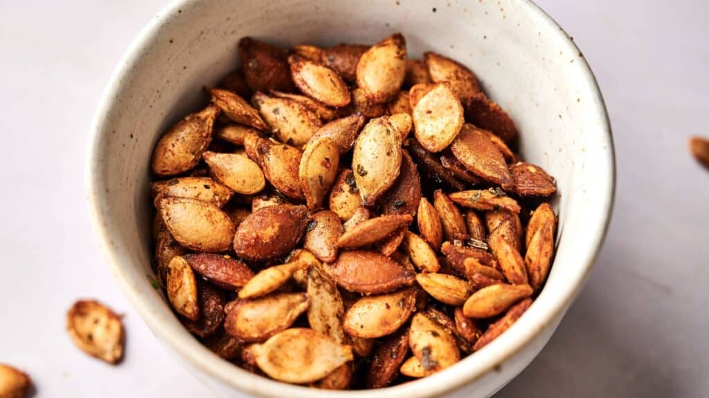 A white bowl filled with roasted, seasoned pumpkin seeds on a light surface.