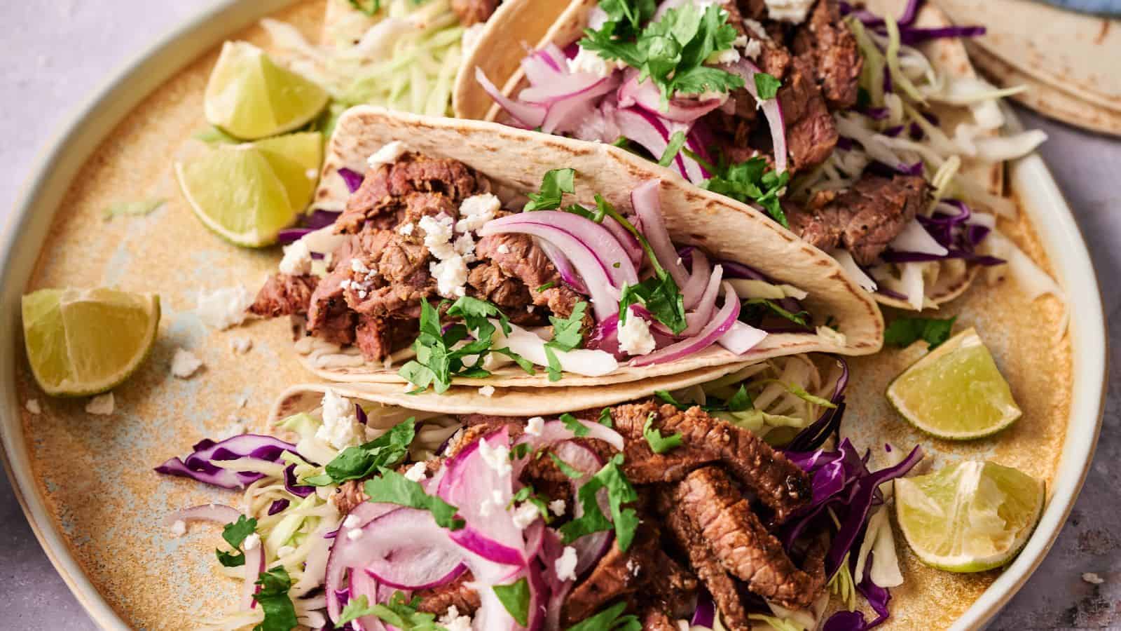 Two beef tacos on a plate, topped with sliced red onions, shredded cabbage, cilantro, and crumbled cheese, surrounded by lime wedges.