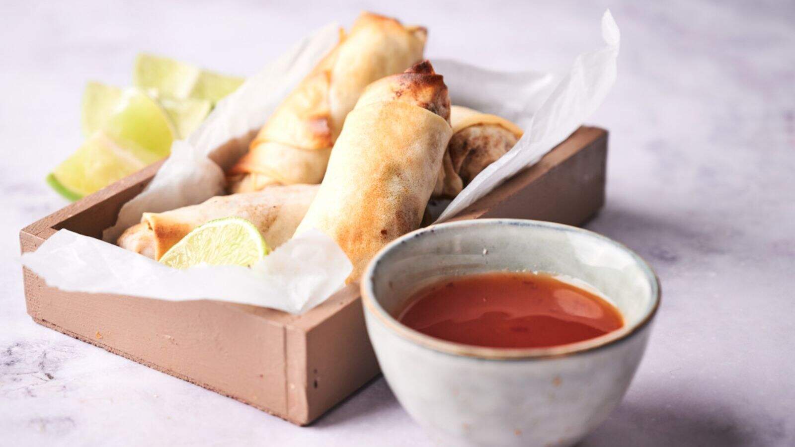 A wooden tray with spring rolls and lime wedges lined with parchment paper sits next to a small bowl of dipping sauce on a light surface.