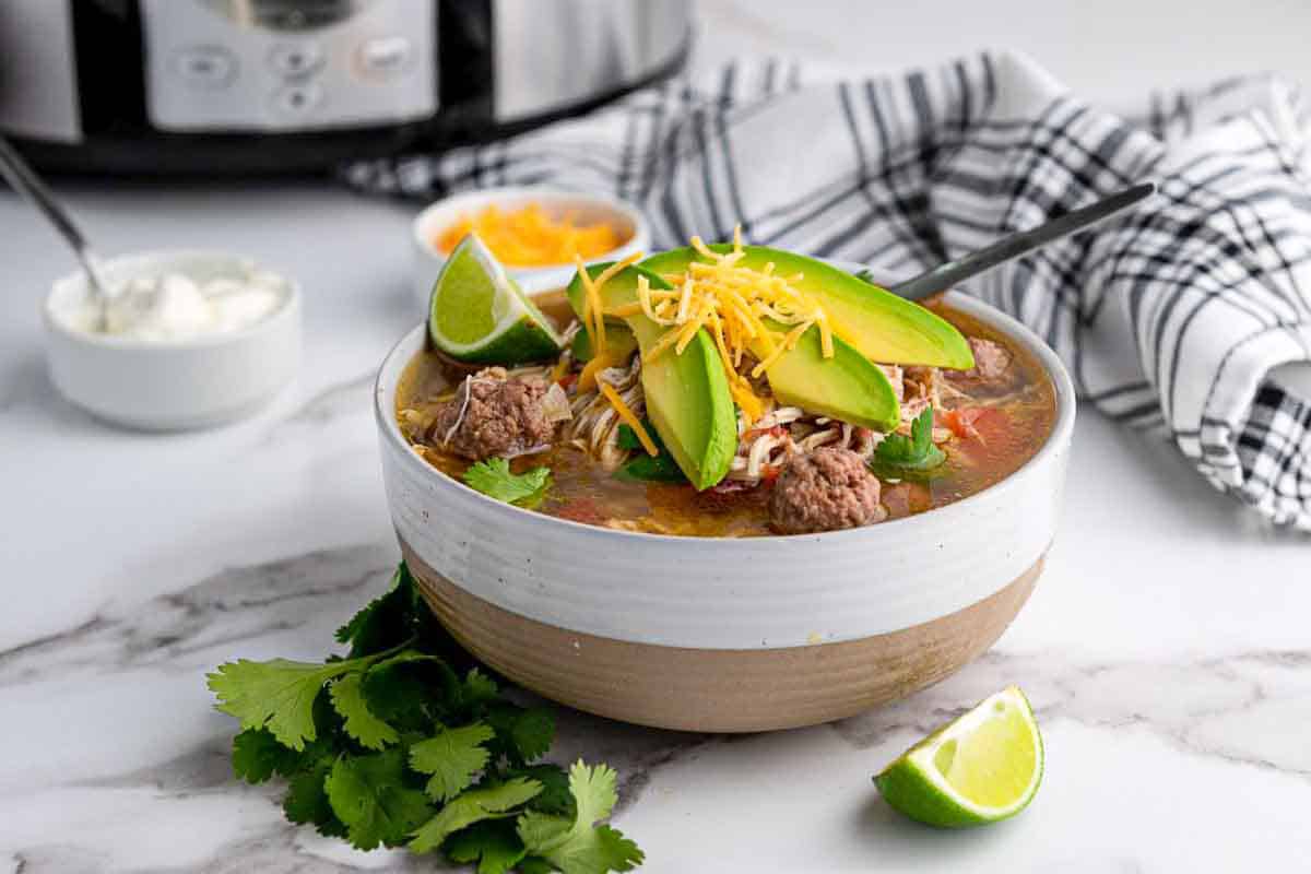 A bowl of Mexican taco soup topped with avocado slices, shredded cheese, and cilantro sits on a marble counter near a lime wedge, cilantro leaves, sour cream, and a slow cooker.