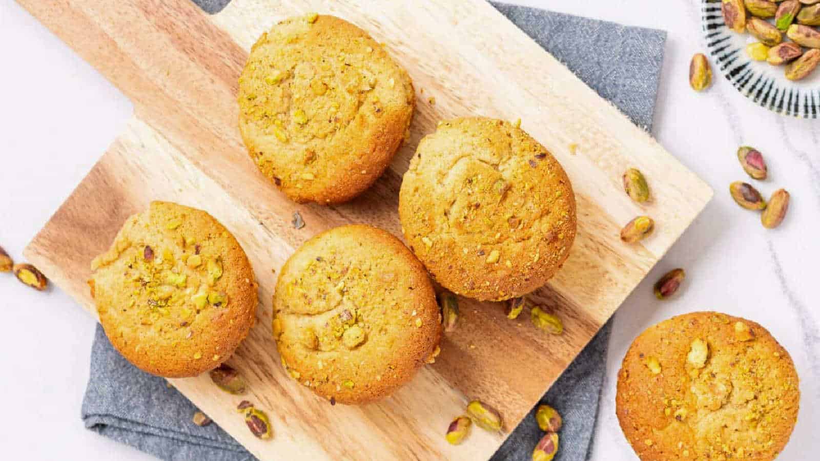 Top view of four pistachio muffins on a wooden board, with one muffin placed separately and pistachios in the background.