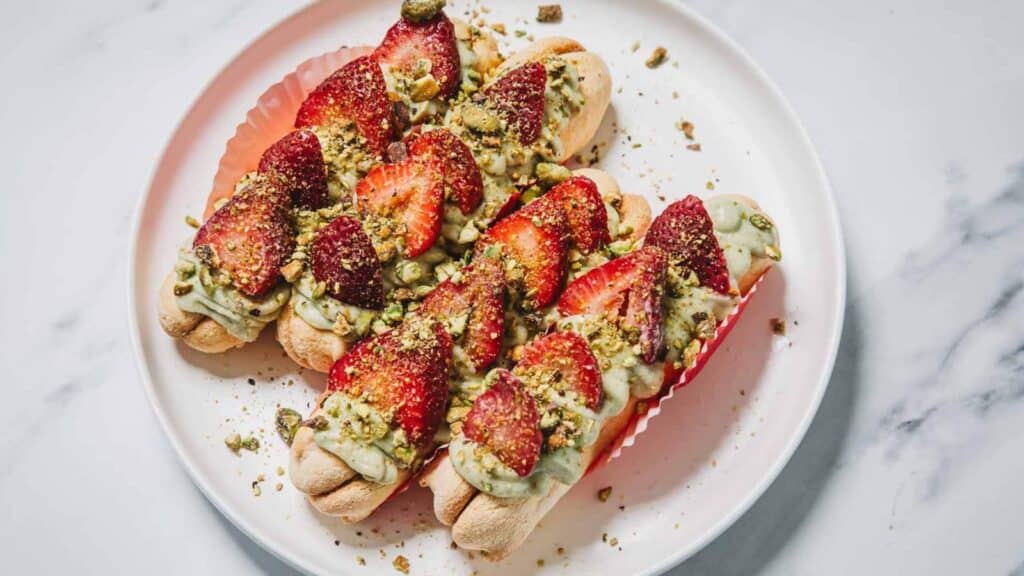 Three ladyfinger pastries topped with green cream, sliced strawberries, and chopped pistachios on a white plate.