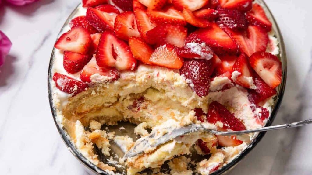 A dish of strawberry tiramisu with a spoon and a portion already served, topped with fresh sliced strawberries.