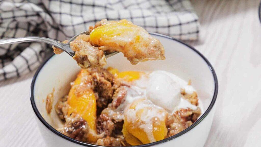 A bowl of peach cobbler topped with melting vanilla ice cream, with a spoonful being lifted, and a checkered cloth in the background.
