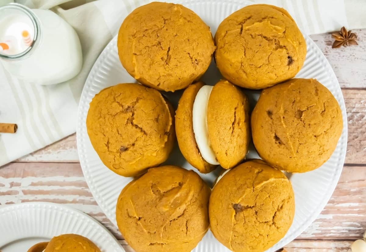 A plate of seven pumpkin whoopie pies with creamy filling is arranged on a wooden table. A glass of milk with straws sits nearby, evoking a cozy autumn feel.