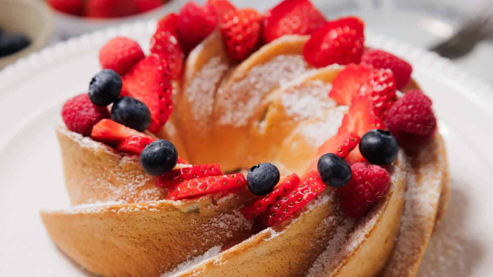 A Bundt cake topped with powdered sugar, fresh strawberries, blueberries, and raspberries sits on a white plate.