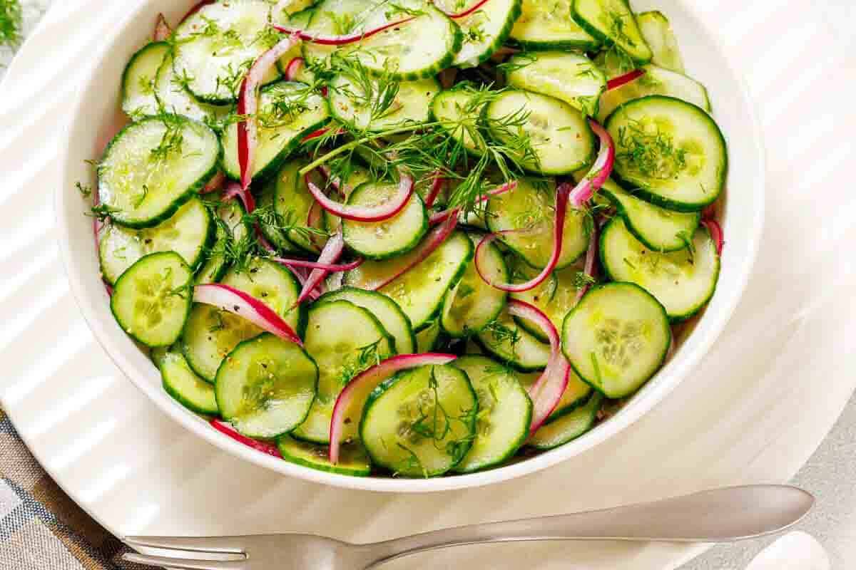 A white bowl filled with sliced cucumbers, red onions, and fresh dill, placed on a white plate with a fork and spoon beside it.