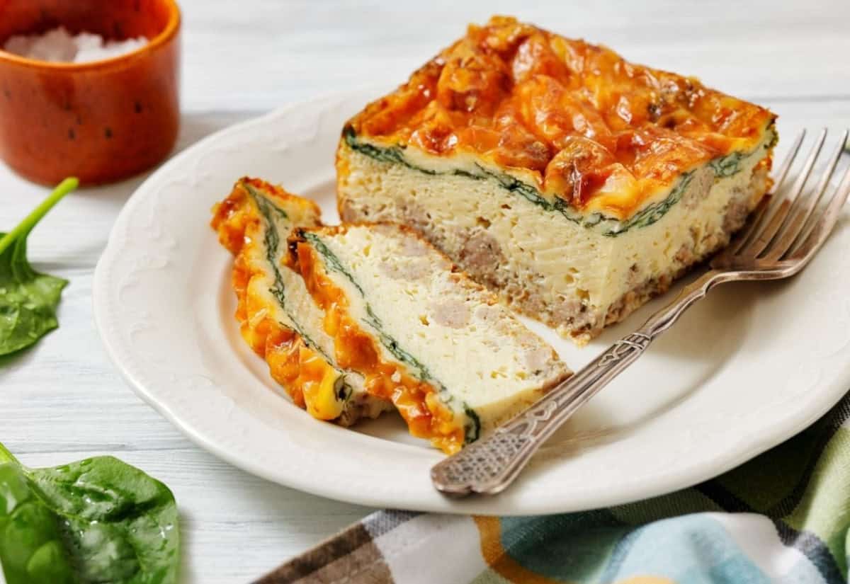A savory breakfast casserole slice with cheese, spinach, and sausage on a white plate, accompanied by a fork, fresh spinach leaves, and a salt bowl.