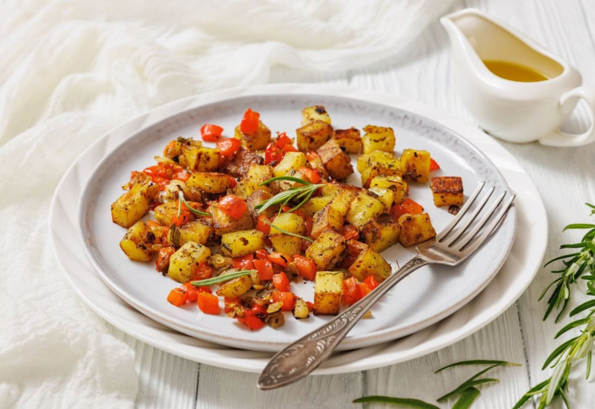Plate of roasted potatoes with red bell peppers and herbs on a white dish, accompanied by a vintage fork. Olive oil in a gravy boat is in the background.