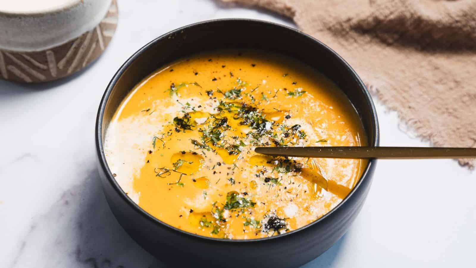 A bowl of creamy orange soup topped with herbs, black pepper, and a drizzle of oil, with a spoon resting inside.